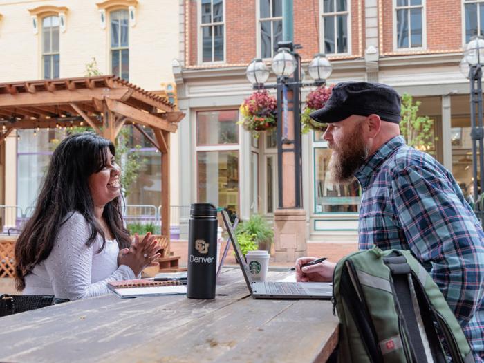 Two CU Denver students sitting across from each other at a table with notebooks and laptops while talking