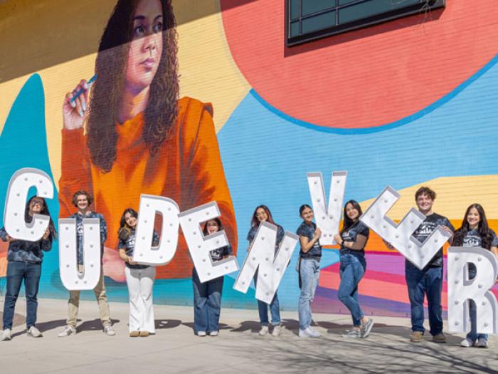 Students holding lighted letters spelling CU Denver in front of on-campus mural