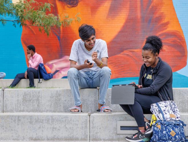 CU Denver students sitting on steps on sitting on steps in front of mural both looking at something on a tablet
