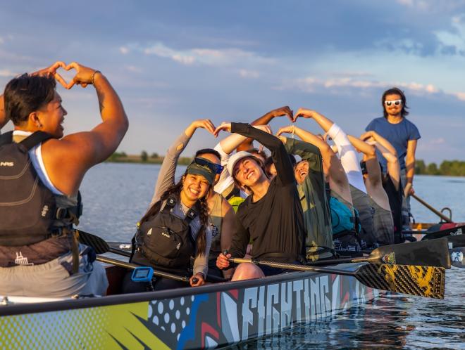 A team of students on a long paddle boat make heart shapes with their hands. 
