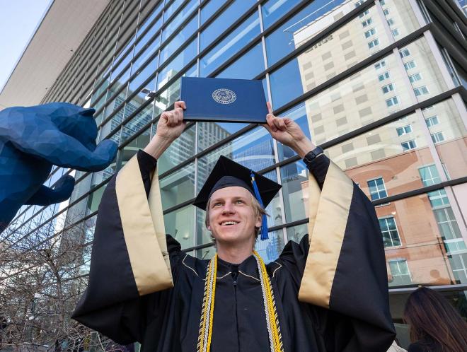 Graduate in regalia holding up diploma cover at commencement