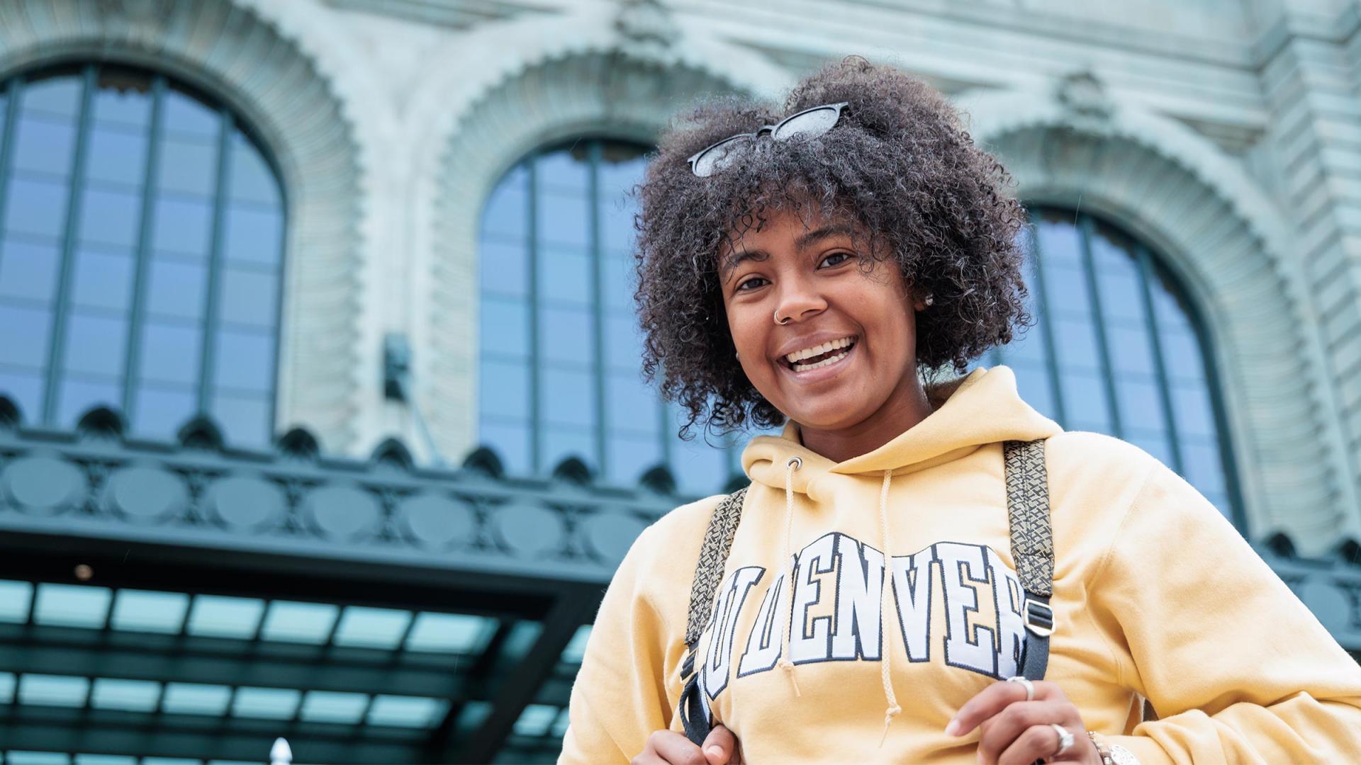 CU Denver student smiling down at building wearing a CU Denver gold hoodie