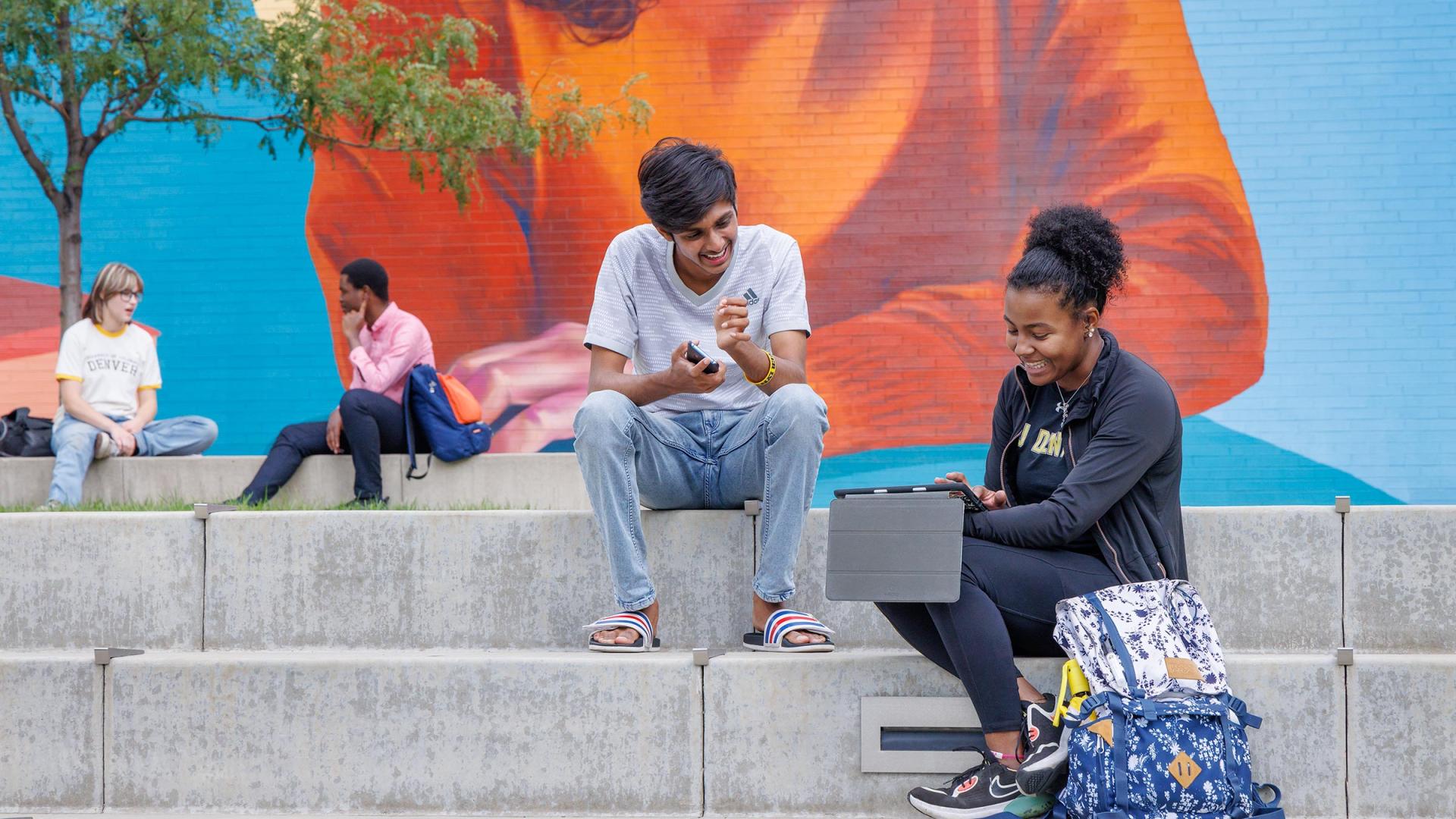CU Denver students sitting on steps on sitting on steps in front of mural both looking at something on a tablet