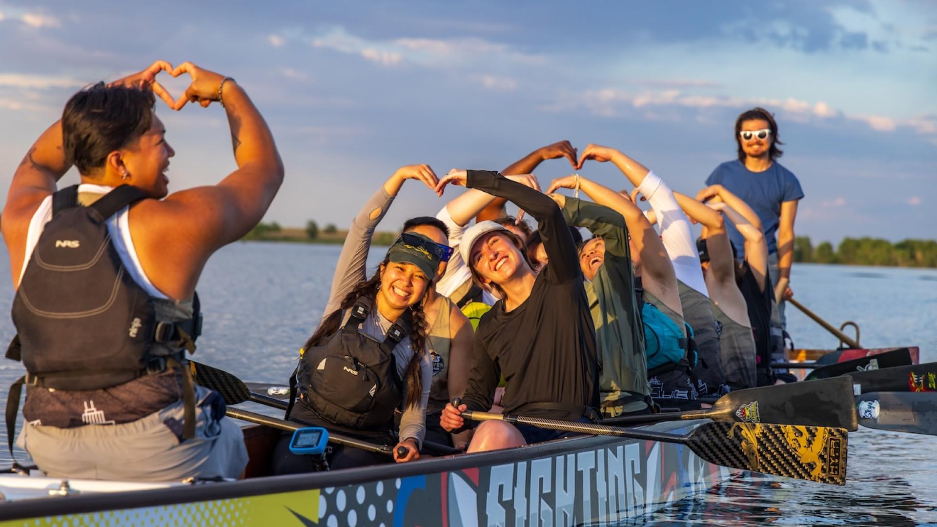 A team of students on a long paddle boat make heart shapes with their hands. 