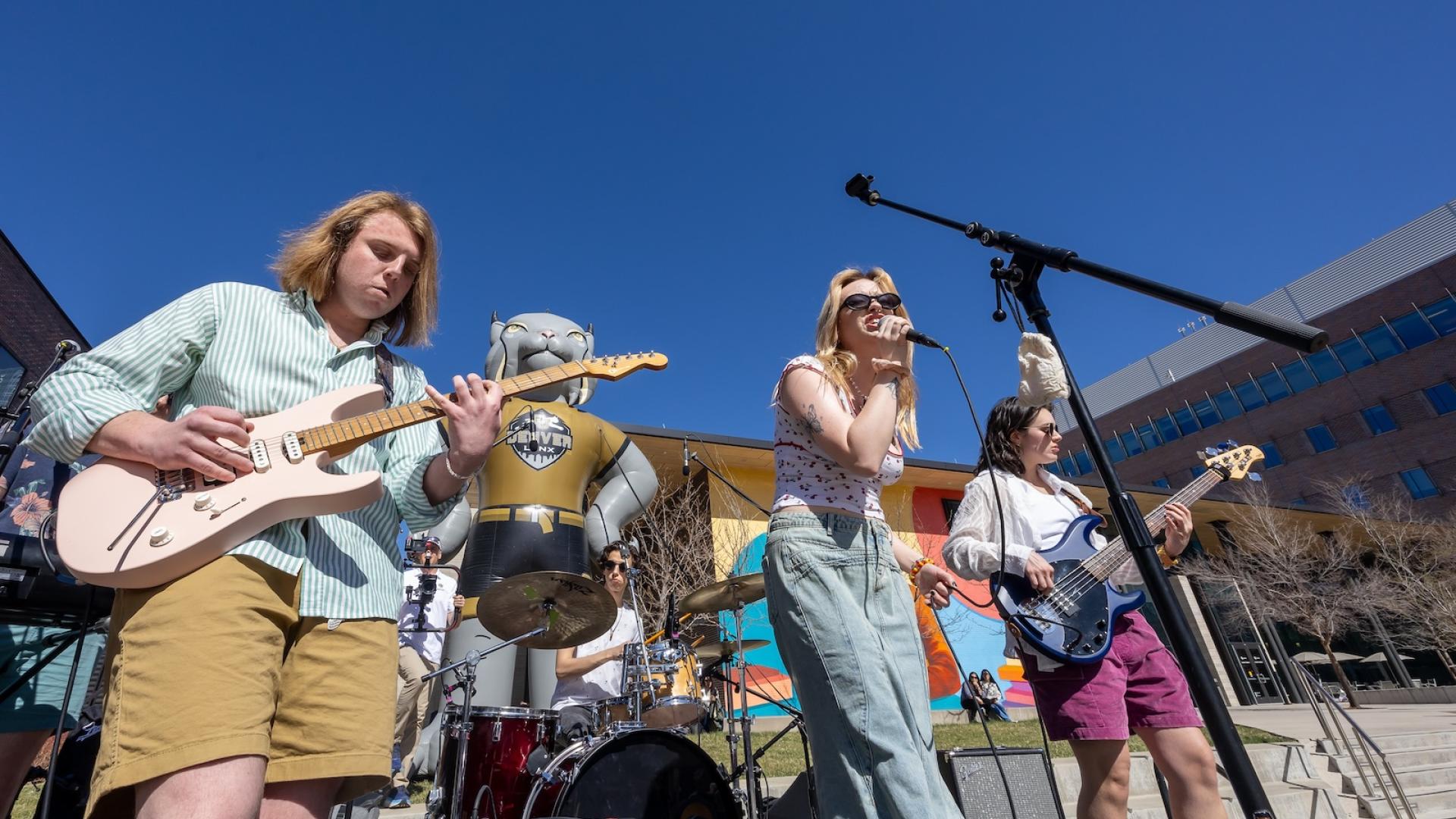 Students play music in a rock band with a large inflatable Milo the Lynx behind them.