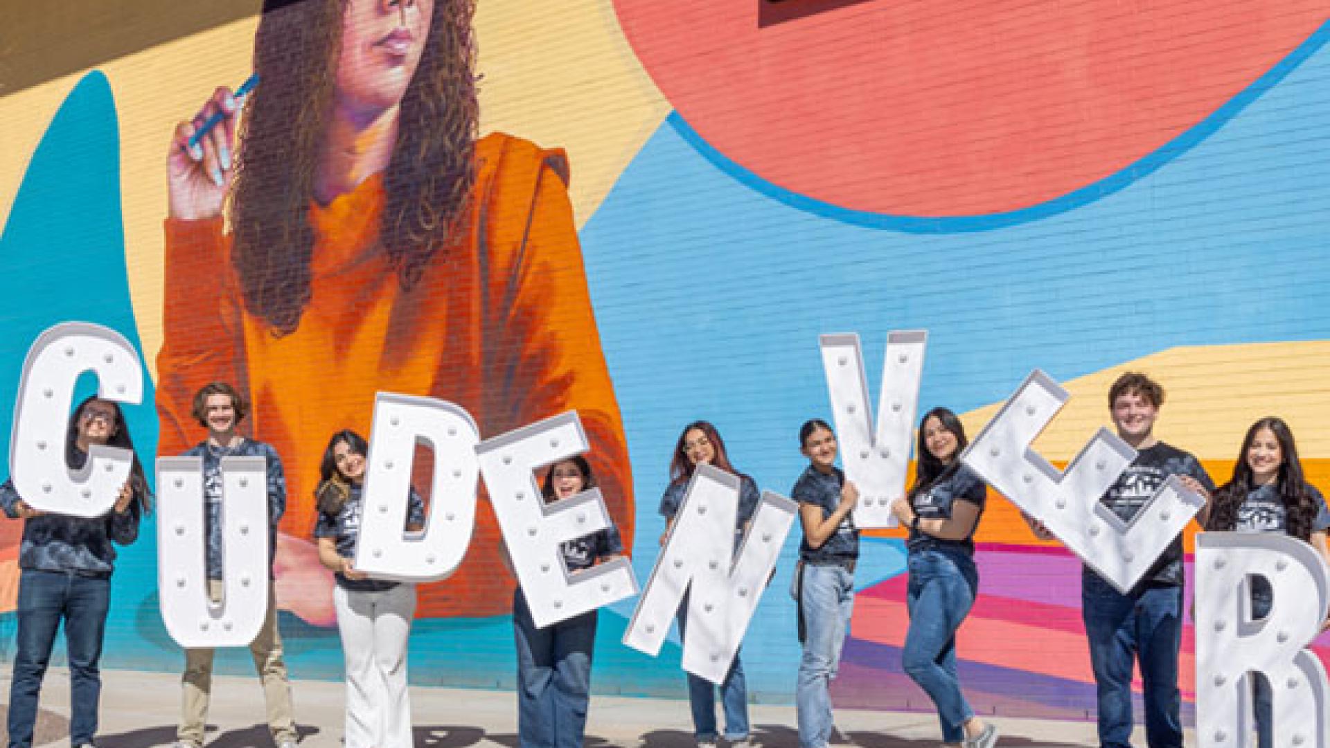Students holding lighted letters spelling CU Denver in front of on-campus mural