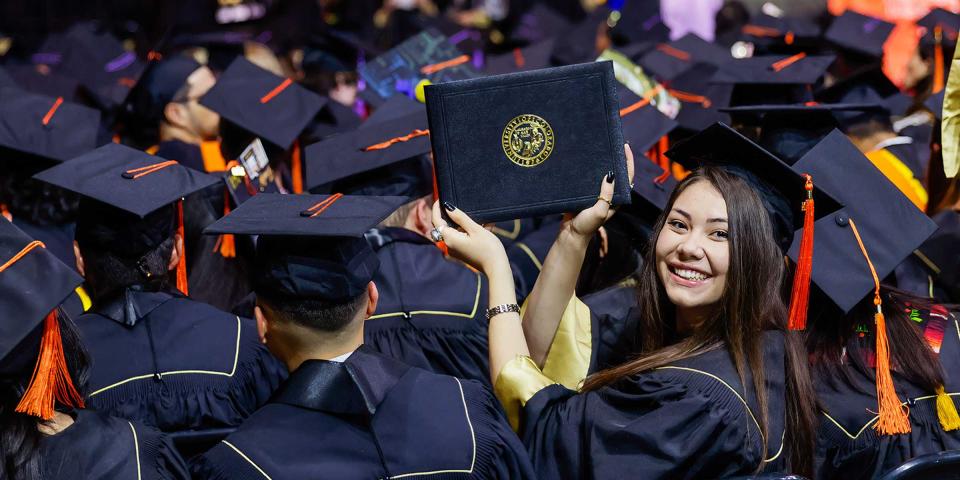 CU Denver graduate turned around facing the camera holding up diploma cover at commencement