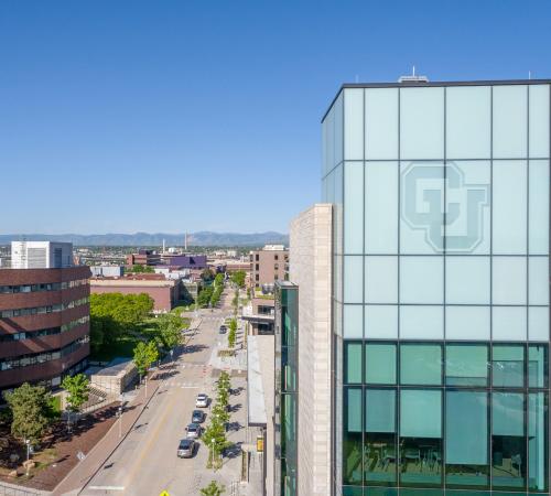 CU Denver building with large glass windows stands along a city street, with tree-lined sidewalks, surrounding campus buildings, and mountains visible in the distance under a clear blue sky.