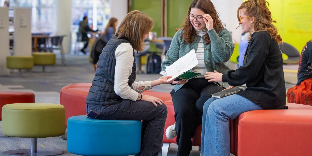 Three students sit together on colorful round seats, reviewing papers and having a discussion while other students work in the background.