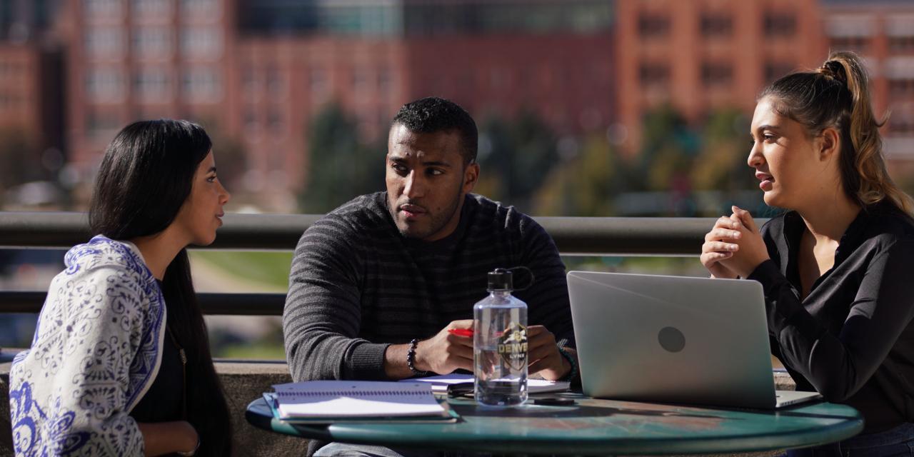 Three University of Colorado Denver students sitting at an outdoor table on an elevated patio, with city buildings in the background, engaged in conversation with notebooks and a laptop on the table.
