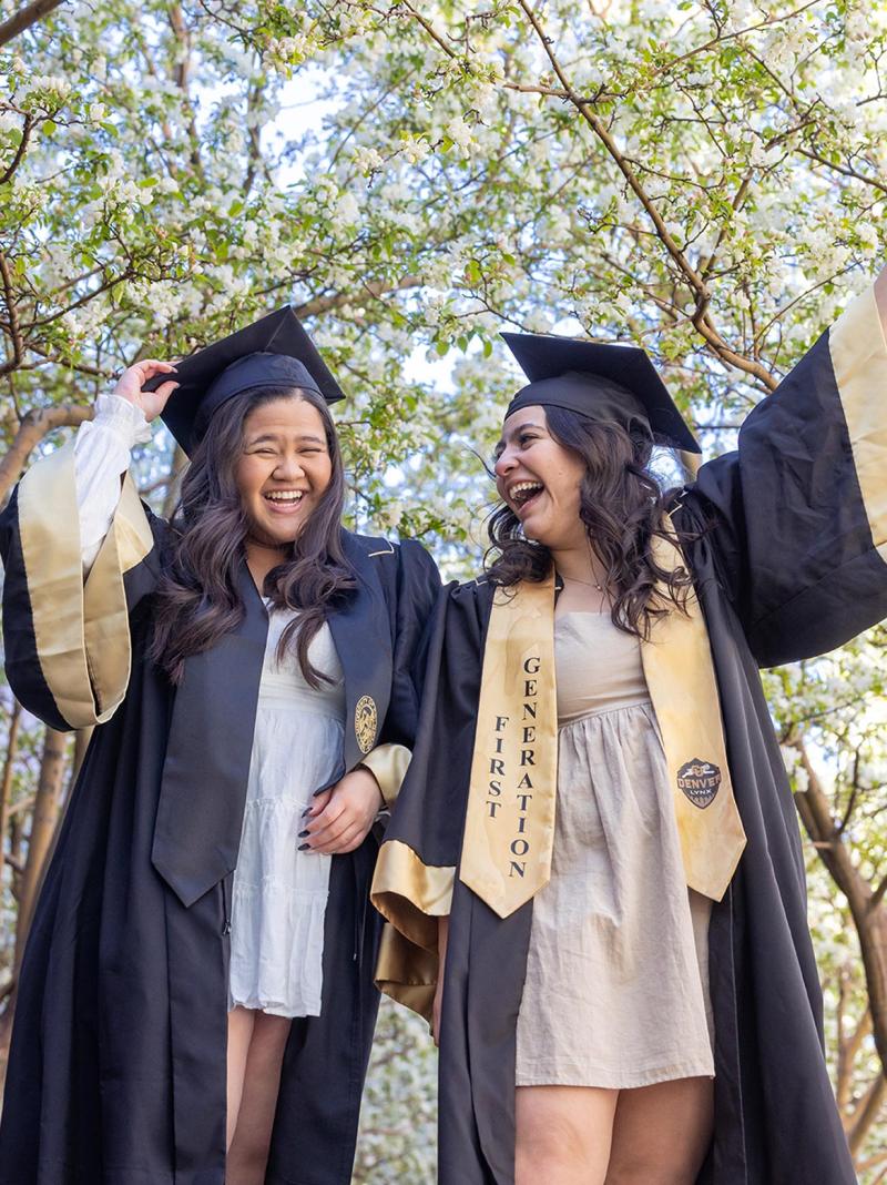 Two CU Denver graduates in regalia with First Generation stole standing in front of trees