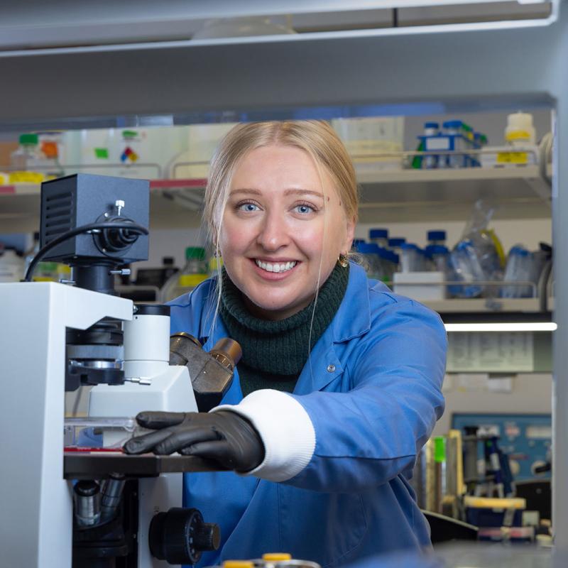 CU Denver student in lab smiling at camera while wearing gloves and using microscope 