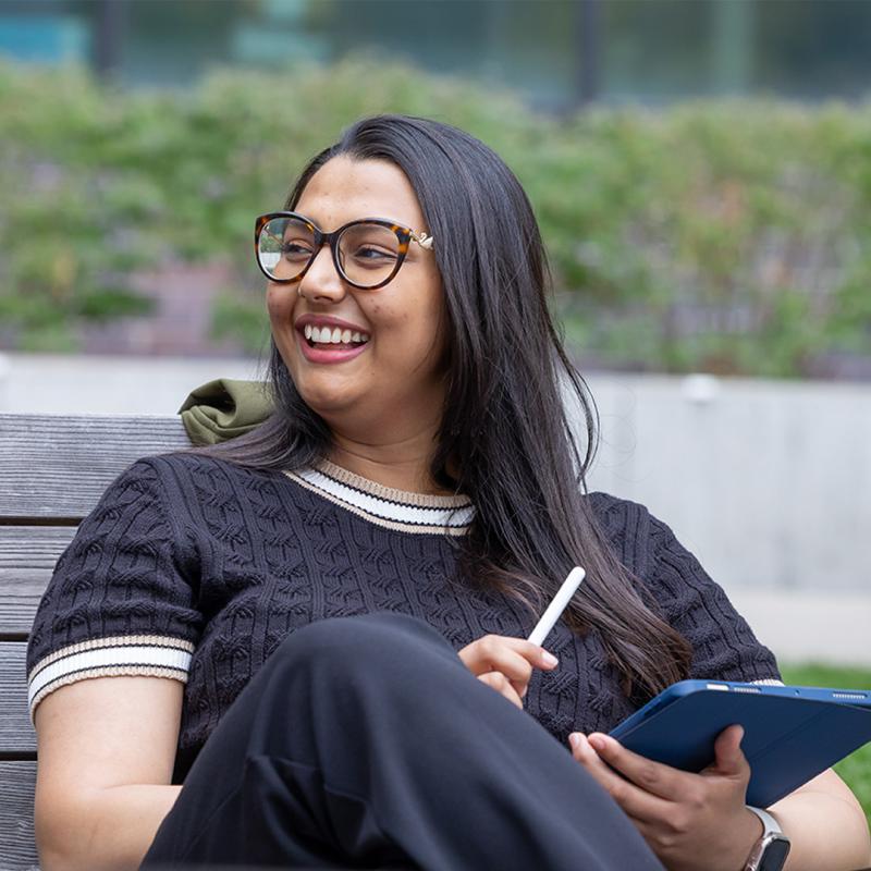 Cu Denver student smiling and looking away from camera while sitting in chair outside with tablet in hand