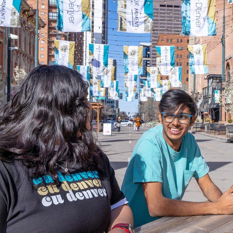 Two students sitting at picnic bench in Larimer Square smiling at each other