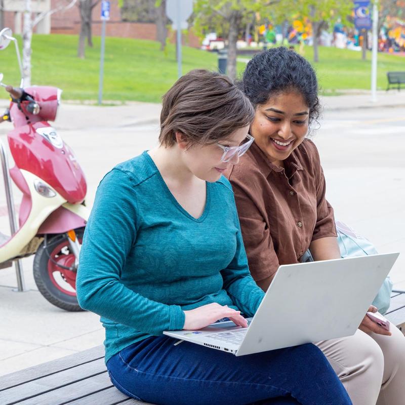 Two students sitting on bench outside looking at laptop