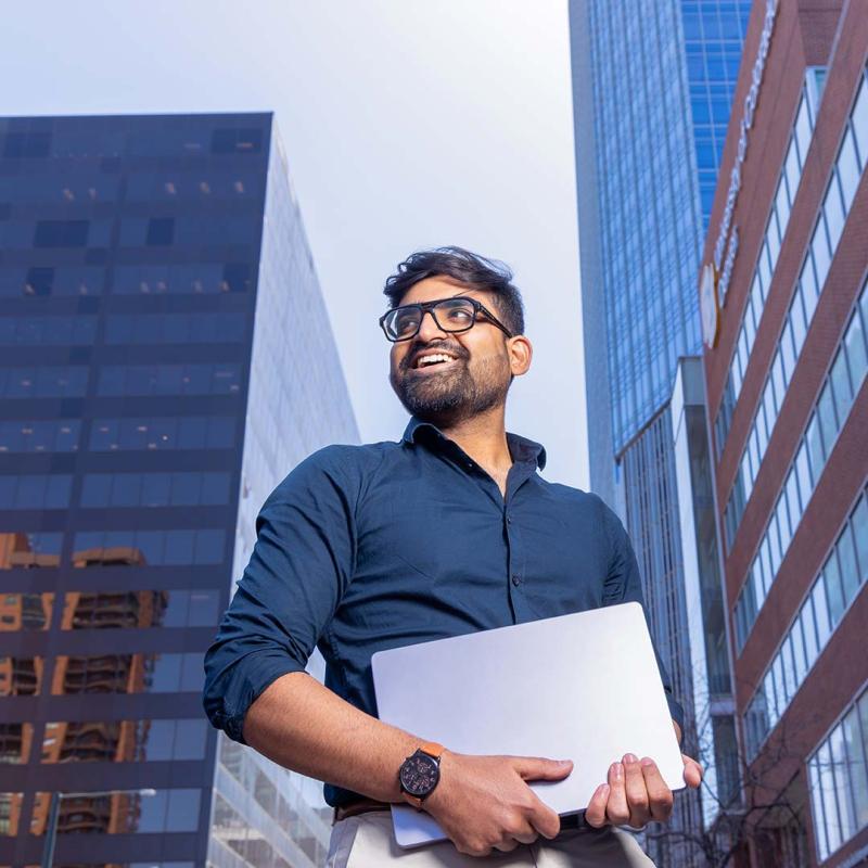 CU Denver alum holding laptop standing in front of buildings in Denver, Colorado