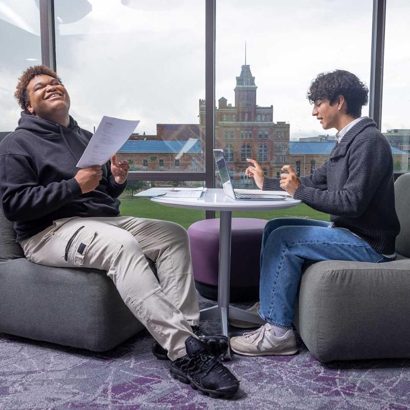 Two students sitting at table in the Student Center with the Tivoli building visible through the window behind them