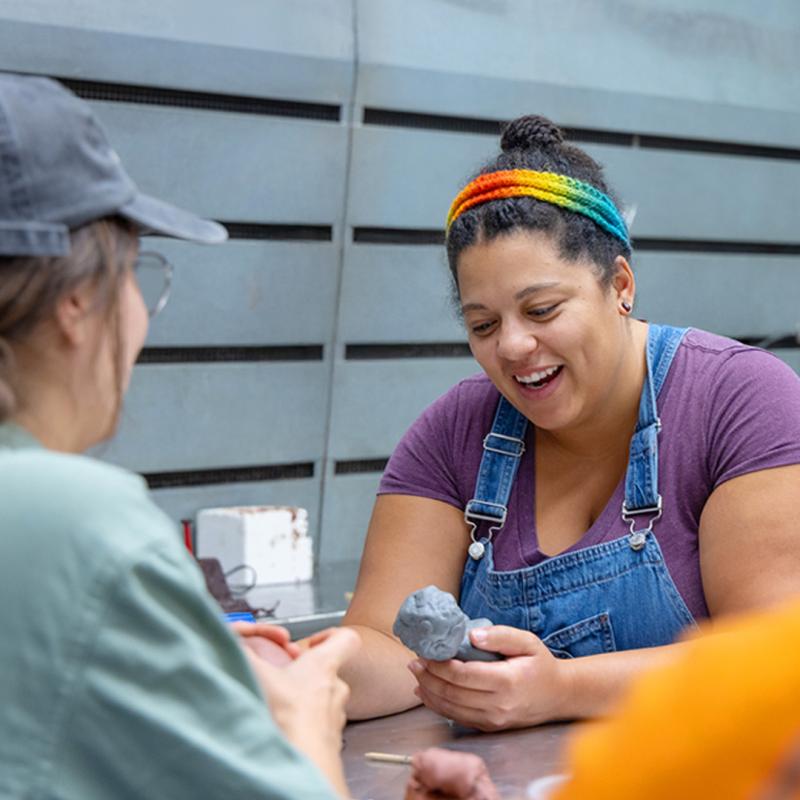 Student wearing purple shirt with jean overalls and rainbow knit headband working with clay in class