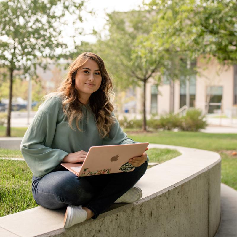 CU Denver student sitting on short wall while working on a laptop