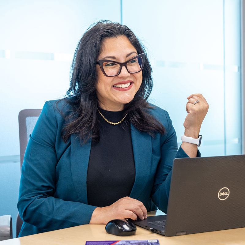 A woman sitting at table wearing blue blazer and black glasses with open laptop talking to someone out of frame and smiling